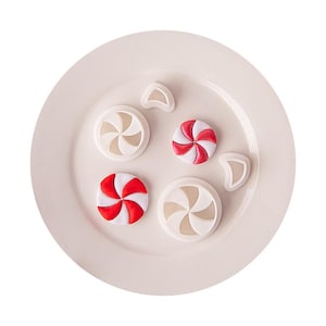 May include: Two round white plastic cookie cutters with a spiral swirl pattern, two finished round peppermint candies with red and white stripes, and two small white crescent-shaped plastic cutters arranged on a plain light-colored plate.