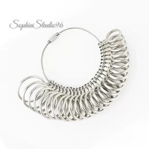 Peut inclure: Un ensemble de baguiers argentés avec plusieurs tailles de bagues attachées à un câble métallique flexible. L'ensemble est conçu pour mesurer avec précision les tailles de bagues. Le texte "Sophia Studio96" est dans le coin supérieur gauche.
