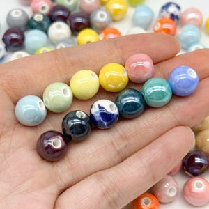 May include: A collection of smooth, round ceramic beads in a variety of glossy colours including pastel pink, bright yellow, light blue, teal, dark blue, and white with blue patterns, each featuring a central hole for stringing and crafting.