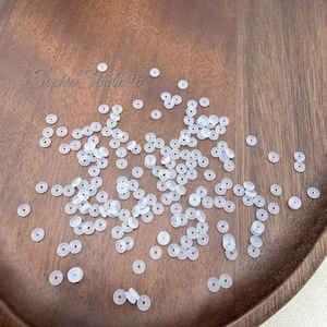 May include: A collection of small, translucent, circular beads with a central hole, scattered across a wooden surface. The beads are clear and matte in appearance. The text "SophiaStudio96" is visible in a cursive font on the wood surface.