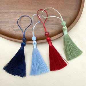 Puede incluir: Cuatro borlas decorativas en azul marino, azul claro, rojo y verde salvia. Cada borla cuenta con un lazo de cordón a juego, una cuenta redonda en la parte superior y una tapa cilíndrica que asegura los hilos finos y rectos.
