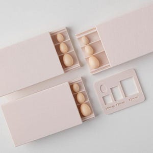 May include: Three light pink sliding storage boxes containing small, smooth, oval-shaped beige beads in graduated sizes. A matching light pink plastic sizing template is included, featuring cutouts labelled 10mm, 12mm, and 15mm.
