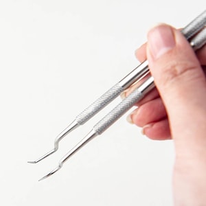 Pode incluir: Duas ferramentas de metal prateado tipo sonda dentária com cabos texturizados e serrilhados para aderência. Cada instrumento possui uma haste fina e lisa que termina numa ponta angulada distinta, concebida para tarefas de precisão e detalhe.
