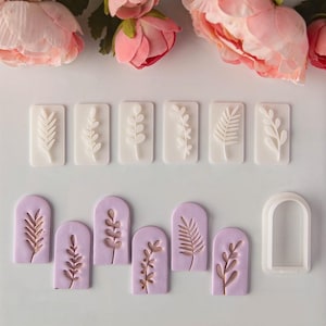 Könnte beinhalten: Ein Set aus sechs rechteckigen weißen Stempeln mit verschiedenen botanischen Blattmustern, sechs bogenförmigen hellvioletten Tonstücken mit geprägten Blattdesigns und einem weißen bogenförmigen Kunststoffausstecher für Ton oder Teig.
