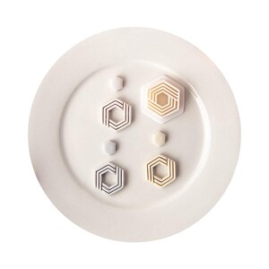 Könnte beinhalten: Eine Sammlung von vier sechseckigen Ausstechformen und zwei kleinen sechseckigen Stempeln auf einem weißen Teller. Die Ausstecher zeigen komplexe geometrische Linienmuster in den Farben Weiß, Creme und Hellgrau innerhalb ihrer Rahmen.