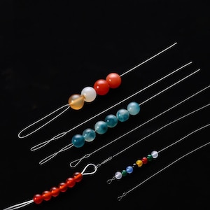 Pode incluir: Variedade de contas coloridas de pedras preciosas enfiadas em fios finos e transparentes. As contas variam em cores, incluindo vermelho, laranja, branco, azul, verde e amarelo. Os fios são loopados nas extremidades, sugerindo o uso na fabricação de joias ou artesanato.