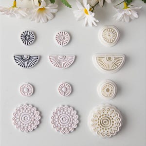 May include: A collection of circular and semi-circular clay cutters and stamped clay shapes arranged in rows. The set includes various sizes of floral and sunburst patterns in white, light pink, and black, featuring intricate radial line details.