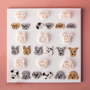 May include: A collection of dog-themed cookie cutters and corresponding molded cookie shapes arranged on a white board. The set includes various canine breeds in shades of tan, gray, white, and black, featuring detailed facial expressions and textures.