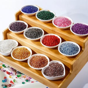 May include: A wooden tiered display holds small white bowls filled with colourful beads. The beads are in various colours, including blue, green, pink, purple, black, red, silver, gold, and brown. Some beads are scattered at the bottom.
