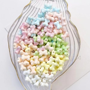 Könnte beinhalten: Eine Ansammlung kleiner, knochenförmiger Perlen in verschiedenen Pastellfarben, darunter Hellblau, zartes Rosa, Hellgrün, Gelb und Weiß. Die Perlen sind in einer durchsichtigen, muschelförmigen Glasschale mit goldfarbenem Rand verstreut.