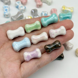 Puede incluir: Una colección de pequeñas cuentas de cerámica en forma de hueso, lisas y brillantes, en varios colores como blanco, azul claro, verde menta, rosa pálido, lavanda y marrón oscuro. Cada cuenta tiene un orificio central y acabado pulido.