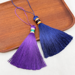 Peut inclure: Deux pompons décoratifs, l'un violet profond et l'autre bleu roi, chacun doté d'un cordon de suspension fin et d'un embout cylindrique orné de rayures horizontales blanches, dorées et noires, fabriqués en fils fins, soyeux et multiples.