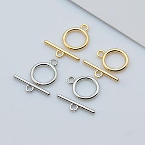 May include: Close-up of gold and silver toggle clasps. The clasps feature a circular ring with a small loop and a straight bar with a small loop. The clasps are used for jewellery making and are displayed on a white surface.