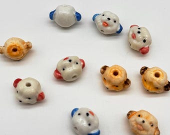 May include: A collection of ten small, hand-painted ceramic beads shaped like animal faces. Each bead features a central hole for stringing, with varied color accents including blue, red, and brown on white or tan glazed ceramic surfaces.