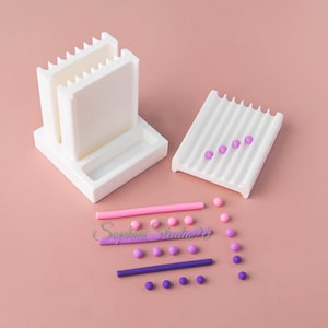 Peut inclure: Un outil extrudeur d'argile en plastique blanc avec un support vertical et un plateau rainuré séparé, accompagné de deux longs bâtonnets d'argile cylindriques rose et violet, et de plusieurs petites perles d'argile sphériques.