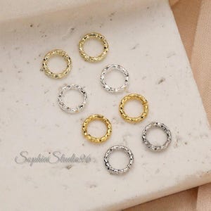 May include: Close-up of several small, textured metal rings in gold and silver tones. The rings are arranged on a light-colored surface. The rings have a textured, faceted design. The image is well-lit, highlighting the metallic sheen.
