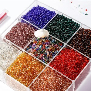 Puede incluir: Un recipiente transparente y dividido lleno de cuentas de semillas de colores. Las cuentas son de varios colores, incluyendo azul, verde, rojo, dorado y marrón. Una concha blanca se encuentra en el centro. Ideal para la fabricación de joyas y manualidades.