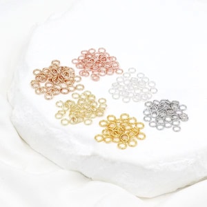 May include: A collection of jump rings in various metallic finishes, including gold, silver, rose gold, and bronze. The jump rings are small, circular, and arranged in separate piles on a white surface. These jewelry-making components are ideal for crafting.