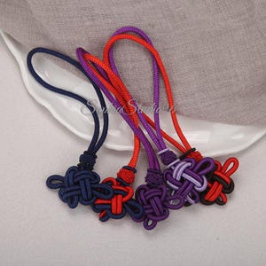 May include: A collection of five decorative cord ornaments featuring intricate knot designs. The set includes individual pieces in solid navy blue, solid red, solid purple, a purple and light purple blend, and a red and black combination.