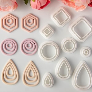 Può includere: Una collezione di vari tagliabiscotti geometrici in tonalità rosa chiaro e bianco. Il set include forme esagonali, circolari, a goccia e quadrate in varie dimensioni, disposte in una griglia su una superficie di colore bianco.