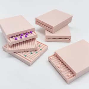 Peut inclure: Un ensemble de rouleaux à perles en plastique rose clair avec des surfaces rainurées, chacun marqué de tailles spécifiques incluant 1,5 mm, 2 mm, 2,5 mm, 3 mm et 4 mm. Les rouleaux contiennent de petites perles sphériques violettes, bleu sarcelle et rose clair.