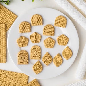 May include: A collection of tan clay shapes with various embossed geometric, floral, and grid patterns arranged on a white plate, accompanied by several white textured rolling pins used for creating these impressions in the clay material.