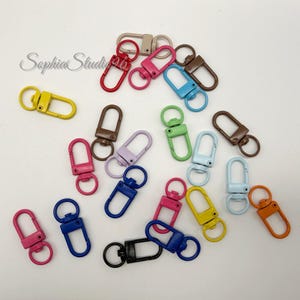 Pode incluir: Uma coleção de fechos lagosta de metal em várias cores, incluindo vermelho, rosa, azul, amarelo, laranja, castanho, preto e lavanda. Cada fecho possui um mecanismo de mola e uma base giratória, espalhados pela imagem.