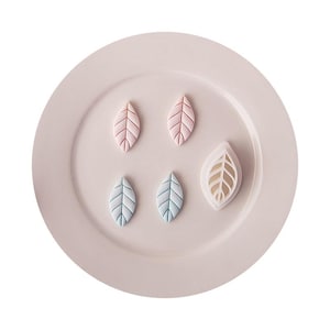 May include: Two small pink leaf-shaped cutouts, two small light blue leaf-shaped cutouts, and one larger white leaf-shaped cookie cutter with a detailed vein pattern, all arranged on a smooth, pale pink circular plate.