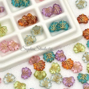 May include: Assortment of colorful leaf-shaped beads in a white tray. The beads are in shades of pink, purple, orange, green, and teal, with gold-colored vein details. The text "Sophia Studio 96" is visible on the tray.