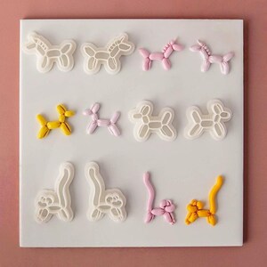 Könnte beinhalten: Eine Sammlung weißer Kunststoff-Ausstechformen in Form von Luftballontieren, darunter Einhörner, Hunde und Katzen, präsentiert neben passenden rosa und gelben Fondant-Figuren auf einem quadratischen weißen Brett.