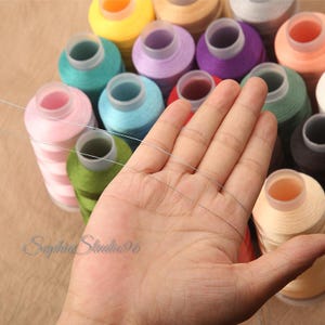 May include: A collection of multi-colored sewing thread spools in shades of yellow, pink, purple, green, blue, and peach, arranged in a cluster. A single thin, light-colored thread is stretched across the foreground. The text SophiaStudio96 is visible.