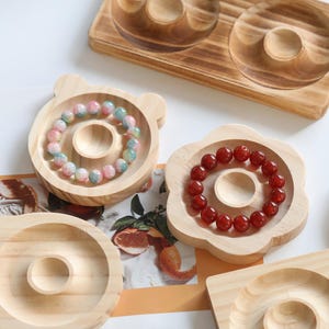 May include: Wooden jewelry display trays in various shapes, including a bear, flower, and round designs. Each tray features a central circular indentation. Bracelets with colorful beads are displayed on the trays. The wood has a natural finish.