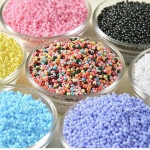 May include: A selection of small glass beads in clear bowls. The beads are in a variety of colours, including pink, yellow, blue, black, white, and a mixed colour bowl. These beads are suitable for jewellery making and crafts.
