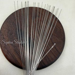 May include: A collection of long, thin, silver-colored metal needles arranged in a fan shape over a circular, dark brown wooden base. The needles are straight with visible eyes at the tips, and the text SophiaStudio96 is printed on the wood.