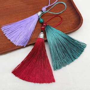 Op de afbeelding: Drie decoratieve kwastjes in lavendel, dieprood en donkergroenblauw. Elk kwastje is voorzien van een zilverkleurige metalen dop, een ophanglus en kleine kleurrijke kralen die in het koord boven de zijdeachtige, fijn gedraaide franjes zitten.