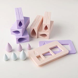 May include: A collection of lavender and peach-colored silicone molds for crafting teardrop shapes, accompanied by six small teardrop-shaped objects in matching colors. The molds feature embossed text indicating sizes in millimeters, including 15mm and 25mm.