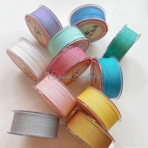 May include: A collection of eleven circular spools of thread in various colors including white, silver, light pink, yellow, gold, light blue, turquoise, green, mint, lavender, and rose. Each spool features a clear plastic rim and a central label.