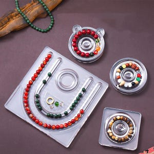 Peut inclure: Un organisateur de perles en plastique transparent avec plusieurs compartiments et des canaux incurvés. L'organisateur contient des perles rouges, vertes et marron, ainsi qu'un collier de perles vertes. La conception comprend des contenants ronds et carrés pour le stockage des perles.