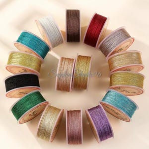 May include: A collection of fifteen circular spools of metallic thread arranged in a circular pattern. The spools feature various colours including white, brown, red, silver, gold, black, green, and purple, each wrapped with thin, shimmering cord.