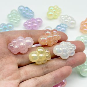 Pode incluir: Uma coleção de pequenas contas em forma de nuvem com acabamento iridescente. As contas são translúcidas e vêm em várias cores pastel, incluindo rosa, amarelo, azul, branco e pêssego. Cada conta apresenta uma textura arredondada.