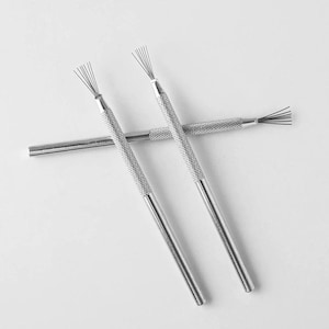 Peut inclure: Deux outils en métal argenté avec des manches texturés et moletés, terminés par de fines brosses en fils métalliques en forme d'éventail. Les outils sont disposés en croix, présentant une finition métallique et un design cylindrique fin.