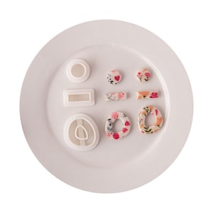 Può includere: Un set di tre tagliapasta in plastica bianca di forma circolare, rettangolare e ovale irregolare, abbinati a tre pezzi di argilla finiti con una base bianca e un colorato motivo floreale nei toni del rosa, arancione e verde.