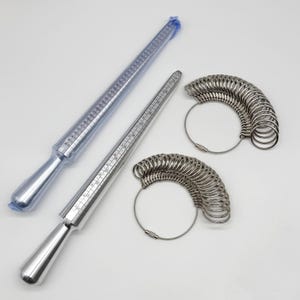 May include: A set of jewelry tools including two silver-colored metal ring mandrels with printed size markings and two sets of graduated metal ring sizers attached to circular wire cables. One mandrel is encased in a clear plastic protective sleeve.