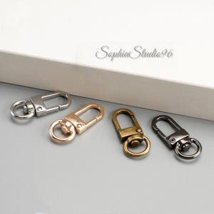 May include: Four metal swivel clasps in silver, gold, antique brass, and gunmetal gray. Each clasp has a D-ring and a spring-loaded gate for secure attachment. The clasps are arranged on a light gray surface.