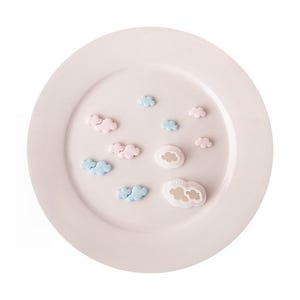 Puede incluir: Una colección de pequeños objetos con forma de nube en colores rosa pastel y azul claro dispuestos sobre un plato rosa pálido. El conjunto incluye varios recortes de nubes individuales y dos moldes de plástico blanco con formas de nubes.