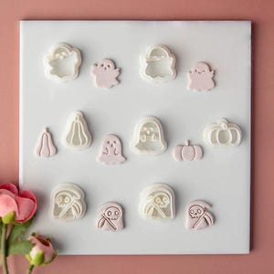May include: A collection of white plastic cookie cutters and corresponding light pink fondant shapes arranged on a white square board. The set includes various Halloween-themed designs such as ghosts, pumpkins, and grim reapers with scythes.