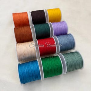 Könnte beinhalten: Eine Sammlung von zwölf Spulen mit farbigem Nylonschnur, die in einem Raster angeordnet sind. Die Spulen zeigen lebendige Farbtöne wie Orange, Dunkelbraun, Gelb, Rost, Dunkelgrün, Lavendel, Beige, Rot, Schieferblau, Hellblau, Grün und Salbei.