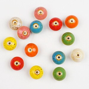 May include: A collection of fourteen round ceramic beads in various colours including red, orange, yellow, green, blue, pink, and cream. Each bead features a smooth, glossy finish with subtle speckled textures and a central hole for threading.