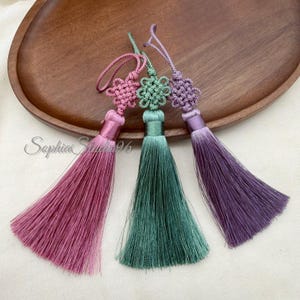 Può includere: Tre nappe decorative in rosa, verde e viola, ciascuna dotata di un cappio superiore annodato e di una finitura in corda liscia simile al raso. Le nappe sono disposte fianco a fianco, mostrando fili lunghi, dritti e sottili che pendono da una fascia centrale.