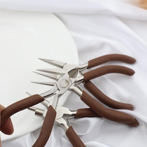 May include: A collection of silver metal jewelry pliers with brown cushioned handles. The pliers are of various types, including needle-nose and side cutters, and are arranged on a white surface.